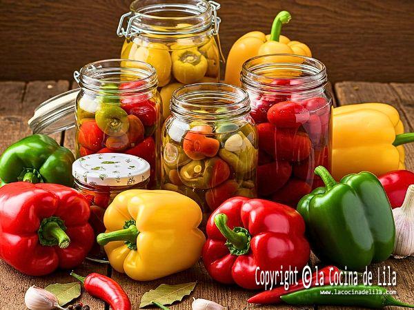 Frascos de vidrio con pimientos de colores (rojos, verdes y amarillos) conservados en aceite y vinagre, acompañados de ingredientes frescos como ajo y hojas de laurel, sobre una mesa rústica.